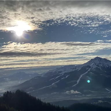 Eine atemberaubende Berglandschaft mit schneebedeckten Gipfeln und einem weiten Tal darunter. Die Sonne scheint durch die Wolken und erzeugt eine schöne Lichtstimmung.