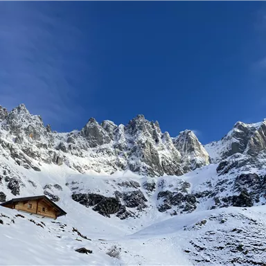 Eine verschneite Berglandschaft mit beeindruckenden Felsen und einem klaren blauen Himmel. Im Vordergrund steht eine kleine Hütte.