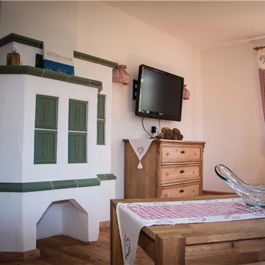 A cozy living room with a traditional tile stove and a television. The wooden table is decorated with a tablecloth and a glass bowl.