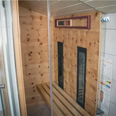 A modern sauna with light wood interior and two heating panels. There is a bench and tiled walls next to a shower cabin.