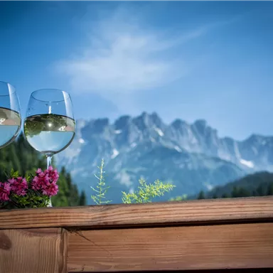 Two glasses of white wine are sitting on a wooden table overlooking majestic mountains. In the foreground, colorful flowers are blooming.