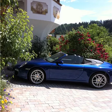 Ein blauer Sportwagen steht auf dem gepflasterten Weg vor einem schönen Haus. Umgeben von grünen Pflanzen und blühenden Blumen.
