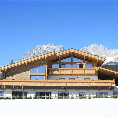 A modern wooden house in the mountains, surrounded by snow and majestic peaks. The clear blue sky completes the idyllic winter landscape.
