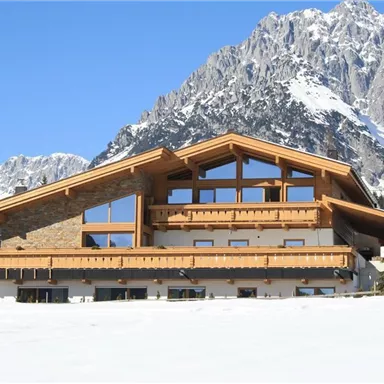 A modern alpine chalet in front of an impressive mountain landscape. The snow-covered ground and the clear sky complete the picturesque scene.