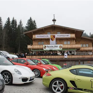 A group of Porsche vehicles is parked in front of a building with a wooden facade. In the background, there is a gathering of the Porsche Club.