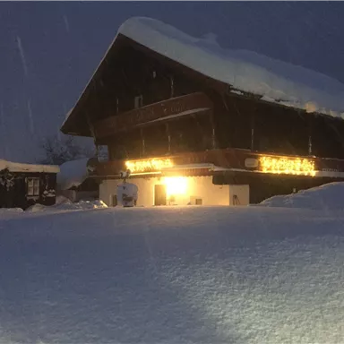 A rustic wooden house in the snow with glowing lights. The surroundings are covered by a thick blanket of snow and the sky is dark.