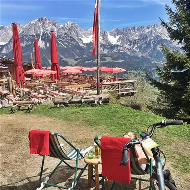 Eine schöne Berglandschaft mit schneebedeckten Gipfeln und einem gemütlichen Biergarten. Liegestühle und ein Mountainbike stehen im Vordergrund.