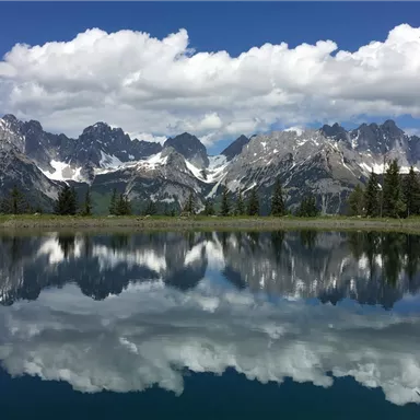 Eine malerische Berglandschaft mit schneebedeckten Gipfeln und einem ruhigen See. Die Wolken spiegeln sich klar im Wasser.