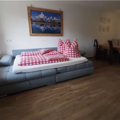 A cozy living space with a large bed and colorful cushions. In the background, a landscape painting hangs on the wall.
