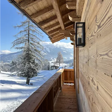 A wooden balcony overlooking a snowy landscape and snow-covered trees. The sky is clear and blue, creating a peaceful winter atmosphere.