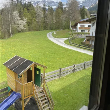 A playhouse with a slide stands on a green meadow. In the background, trees and snow-covered mountains are visible.
