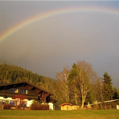 Ein malerisches Haus mit einem Regenbogen im Hintergrund. Die Umgebung ist von Bäumen und grünen Wiesen geprägt.