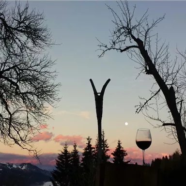 Eine malerische Landschaft mit kahlen Bäumen, einem Glas Wein und einem sanften Sonnenuntergang. Der Himmel ist in sanften Rosa- und Blautönen gefärbt.