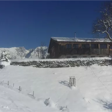 A snow-covered landscape featuring traditional wooden houses. In the background, majestic mountains and a clear blue sky can be seen.