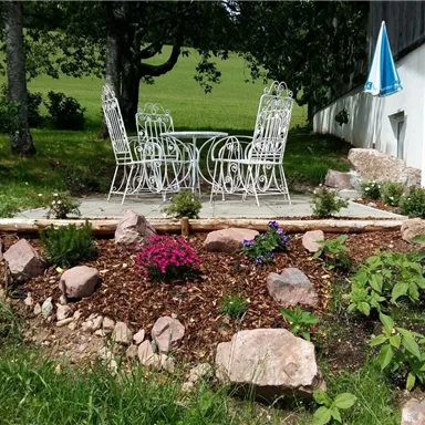 A beautiful garden area with a small seating area and blooming plants. In the background, grasses and trees can be seen.