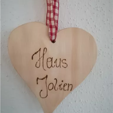Ein herzförmiges Holzschild mit der Aufschrift "Haus Jolien". Es ist mit einem roten karierten Band zum Aufhängen versehen.