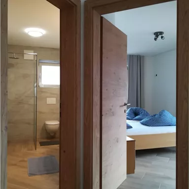 Ein modern eingerichteter Raum mit zwei Türen. Auf der linken Seite ist ein Bad mit Dusche, auf der rechten Seite ein Schlafzimmer mit einem Bett und Kissen.