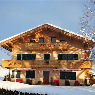 A traditional Alpine house surrounded by snow and mountains. The facade features charming balconies and windows with decorated shutters.