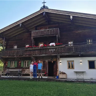 Ein traditionelles, zweigeschossiges Holzhaus in einer malerischen Landschaft. Vor dem Haus stehen drei Personen auf der Veranda.
