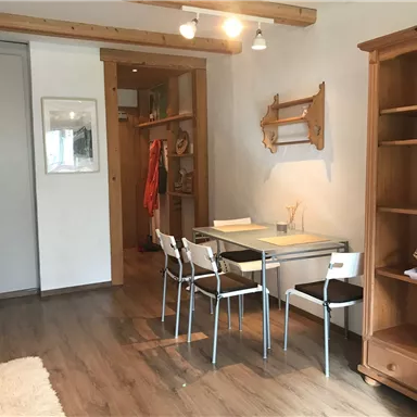 Ein helles Zimmer mit einem Tisch und weißen Stühlen. Im Hintergrund befindet sich ein Regal und ein Kleiderschrank.