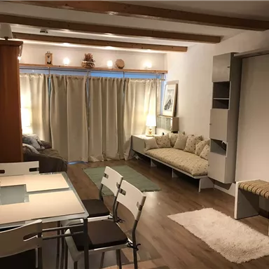 Ein gemütliches Wohnzimmer mit einem Esstisch und Stühlen. Das Zimmer hat helle Vorhänge, einen Sofa-Bereich und warmes Licht.