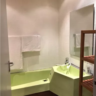 Ein modernes Badezimmer mit einer grünen Badewanne und einem großen Spiegel. Im Raum befinden sich weiße Handtücher und ein Holzregal.