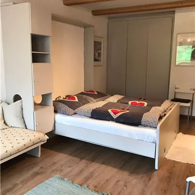 Ein gemütliches Schlafzimmer mit einem Doppelbett und bunten Bettdecken. Im Raum befinden sich ein Regal, eine Couch und ein Tisch mit Stühlen.
