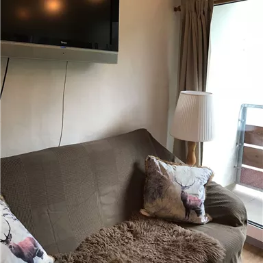 Ein gemütliches Wohnzimmer mit einem braunen Sofa und dekorativen Kissen. Ein Fernseher hängt an der Wand und es gibt eine Lampe neben einem Fenster.