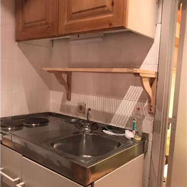 Eine kleine Küchenzeile mit einem Doppelspülbecken und zwei Kochplatten. Über der Arbeitsfläche befindet sich ein Regal aus Holz.