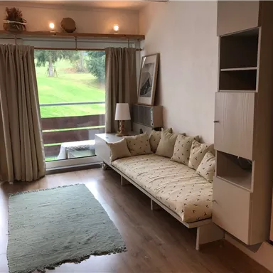 Ein helles Wohnzimmer mit einer gemütlichen Sitzbank und einem großen Fenster. Draußen ist eine grüne Wiese sichtbar.
