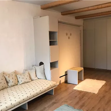 Ein gemütlicher Wohnraum mit einem bequemen Sofa und hellen Wänden. Er enthält einen kleinen Tisch und einen Kleiderschrank sowie einen weichen Teppich.
