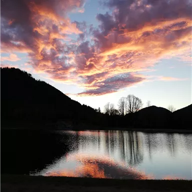 Ein ruhiger See, umgeben von Bergen, spiegelt einen wunderschönen Sonnenuntergang mit bunten Wolken wider. Die Atmosphäre ist friedlich und malerisch.
