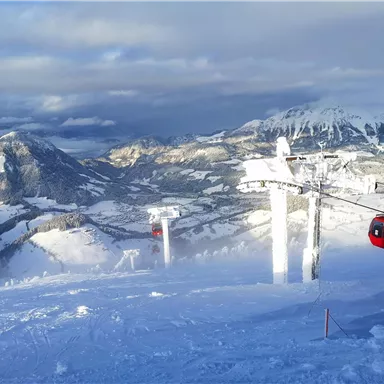 A snowy mountain landscape with ski lifts and peaks in the background. The sky is partly cloudy, and the view is breathtaking.