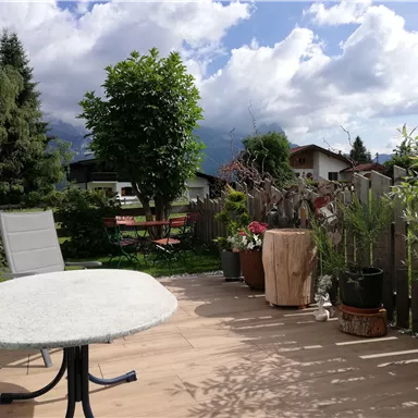Eine Terrasse mit einem Tisch und einem Stuhl, umgeben von Pflanzen und Bäumen. Der Himmel ist teilweise bewölkt mit etwas Sonnenschein.