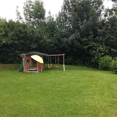 Eine Spielwiese mit einem kleinen Spielhaus und einer Schaukel steht im Vordergrund. Im Hintergrund sind hohe Hecken und Bäume zu sehen.