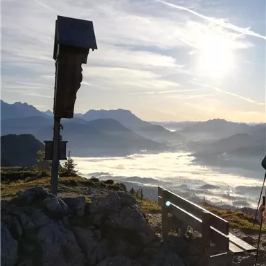 A view of a picturesque mountain landscape with fog in the valley and the sun in the sky. In the foreground, there is a wooden bench and a small wooden structure.