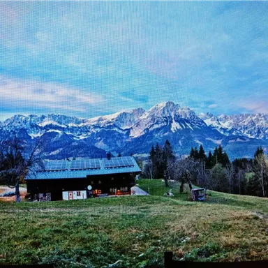 Eine malerische Alpenlandschaft mit schneebedeckten Bergen im Hintergrund. Im Vordergrund steht ein gemütliches Gebäude auf einer grünen Wiese.