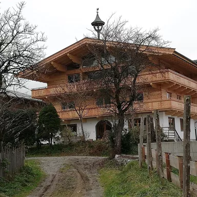 Ein traditionelles Holzhaus mit mehreren Balkonen und einem charakteristischen Dach. Der Garten ist von Bäumen umgeben und ein Schotterweg führt zum Eingang.