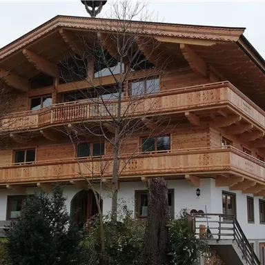 Ein traditionelles Holzhaus mit mehreren Balkonen und großen Fenstern. Rund um das Haus befinden sich Bäume und kleine Sträucher.