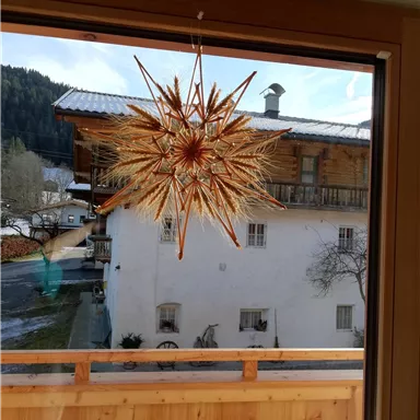Ein schön dekorierter Stern hängt vor einem Fenster. Im Hintergrund ist ein traditionelles Haus und eine malerische Winterlandschaft zu sehen.