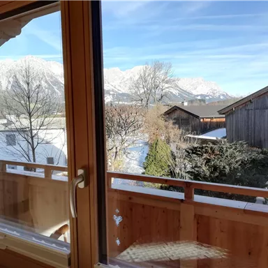 Ein Blick aus einem Holzfenster auf eine winterliche Landschaft. Im Hintergrund sind schneebedeckte Berge und traditionelle Holzhäuser zu sehen.