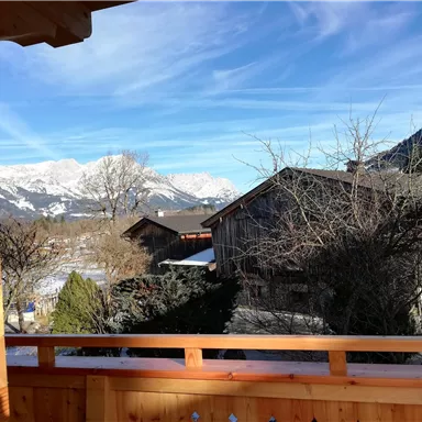 Ein Blick auf die Alpen mit schneebedeckten Bergen und traditionellen Holzhäusern. Der Himmel ist klar und blau mit einigen Wolken.