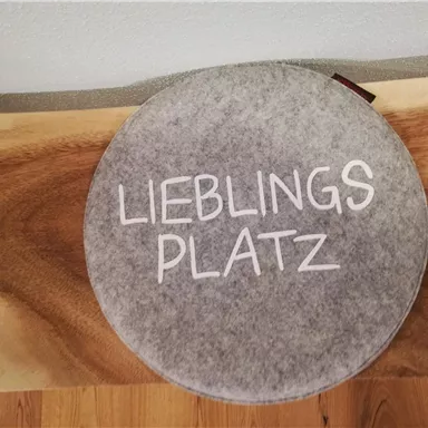 Ein runder grauer Sitzplatz mit dem Aufdruck „LIEBLINGSPLATZ“. Er befindet sich auf einer Holzoberfläche.