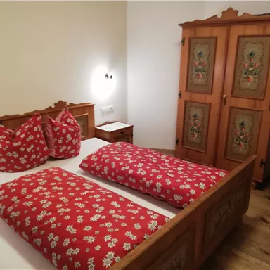 Ein gemütliches Schlafzimmer mit einem Holzbett und roten Kissen. Im Hintergrund steht ein dekorierter Kleiderschrank.