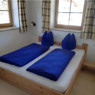 A cozy bedroom with two separate beds and blue bedspreads. The windows allow plenty of daylight in and the walls are painted light.