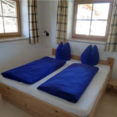 A cozy bedroom with a double bed and blue bedspreads. Light-filled windows and simple wooden furniture give the room a warm atmosphere.