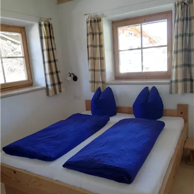 A cozy bedroom with two single beds and blue bedspreads. Large windows provide plenty of daylight.