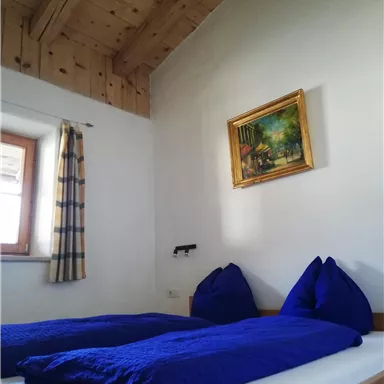 A cozy bedroom with light walls and a wooden ceiling beam. Two blue cushions and a painting on the wall complete the tranquil atmosphere.