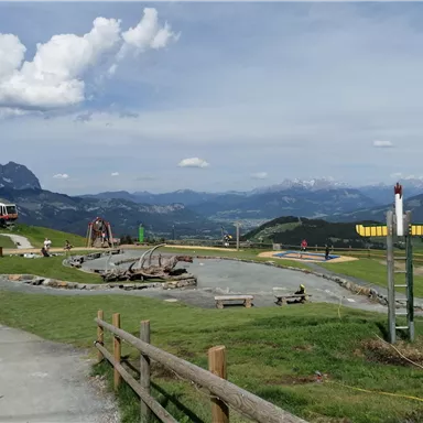 A picturesque mountain landscape with a hiking trail and play equipment. In the background, the mountains and a train can be seen.