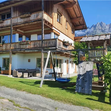 Ein gemütliches Holzhaus mit mehreren Balkonen und einer Terrasse. Im Vordergrund gibt es einen Spielplatz mit einer Schaukel und eine schöne Bergkulisse im Hintergrund.